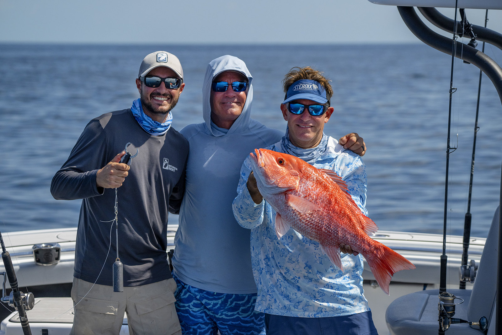 Three anglers stand together on a boat, posing for a photo with thumbs up. One angler is holding a Red snapper.
